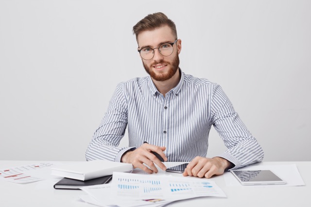 People, career, business and occupation concept. Pleased bearded stylish man with trendy hairdo, wears spectacles, works with modern gadgets and documents, isolated over white studio background